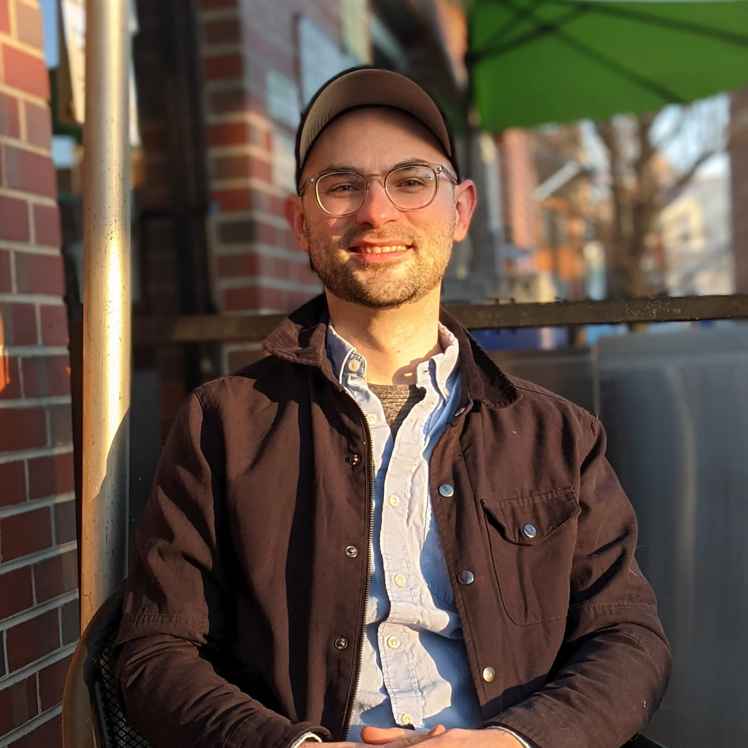 Patrick Morgan, a technical writer in Richmond, Virginia, sitting outside at a local café. He is smiling at the camera, wearing glasses, a blue collared shirt, a dark jacket, and a baseball cap.