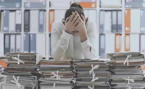 A frustrated office worker leaning on tall piles of paperwork stacked across her desk.